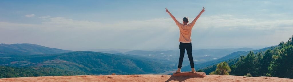 A man raises his arms in triumph on a rocky mountain summit overlooking a vast landscape