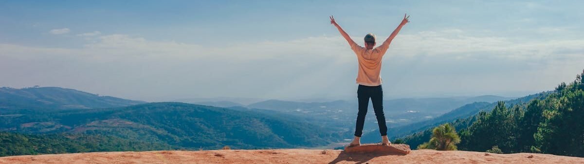 A man raises his arms in triumph on a rocky mountain summit overlooking a vast landscape